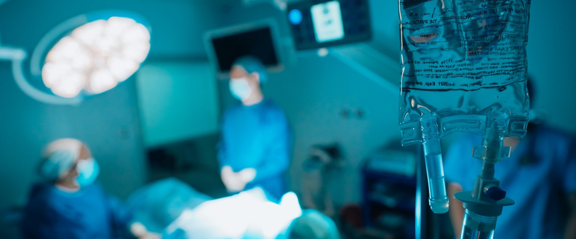 Operating room scene with surgical lighting, medical monitors, and an IV bag in the foreground